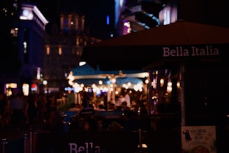 A lively nighttime street scene featuring an outdoor dining area adorned with string lights, with the Bella Italia sign visible on an umbrella. People are gathered, creating a bustling atmosphere filled with warm lighting and shadowy figures. Buildings in the background are dimly lit, adding to the urban ambiance.