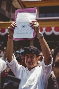 A person is holding up a document with multiple signatures and a stamp, standing among a crowd of people. The individual is dressed in a white shirt and wearing a black headband. The background shows part of a building with red and white banners.