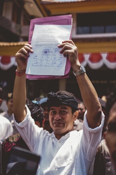 A person is holding up a document with multiple signatures and a stamp, standing among a crowd of people. The individual is dressed in a white shirt and wearing a black headband. The background shows part of a building with red and white banners.