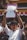 A person is holding up a document with multiple signatures and a stamp, standing among a crowd of people. The individual is dressed in a white shirt and wearing a black headband. The background shows part of a building with red and white banners.