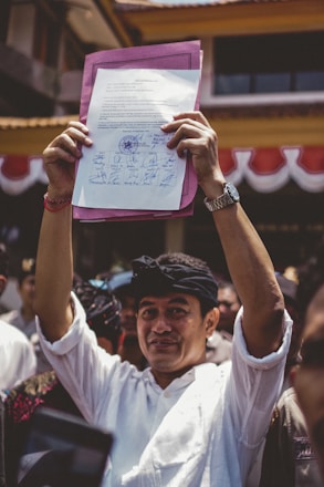 A person is holding up a document with multiple signatures and a stamp, standing among a crowd of people. The individual is dressed in a white shirt and wearing a black headband. The background shows part of a building with red and white banners.