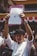 A person is holding up a document with multiple signatures and a stamp, standing among a crowd of people. The individual is dressed in a white shirt and wearing a black headband. The background shows part of a building with red and white banners.