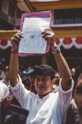 A person is holding up a document with multiple signatures and a stamp, standing among a crowd of people. The individual is dressed in a white shirt and wearing a black headband. The background shows part of a building with red and white banners.