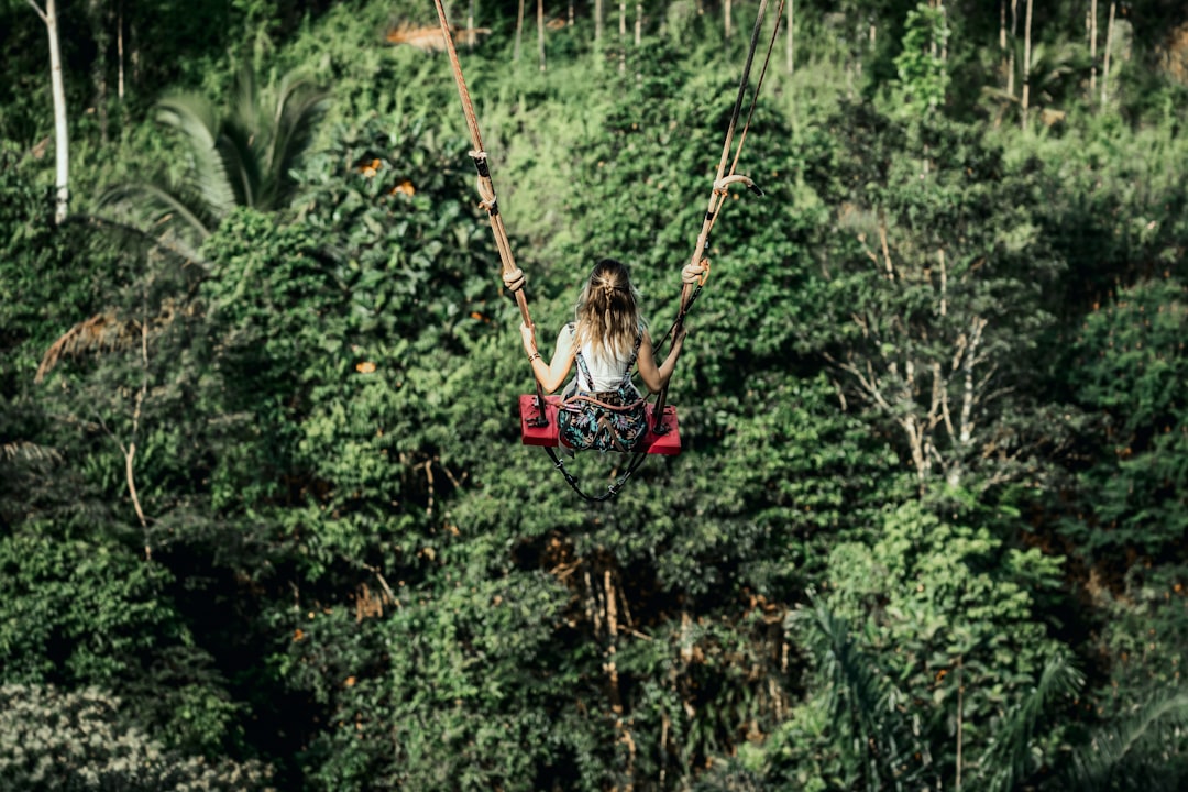 girl in pink jacket riding on swing surrounded by green plants during daytime