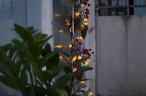A vertical arrangement of holiday decorations featuring pine cones, small globe lights, and red berries. The string of lights is wrapped around a piece of wood, casting a warm glow. Nearby, there are green leaves partially obscuring the view.