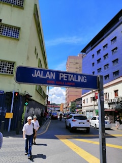 A street scene in an urban area with a street sign reading 'Jalan Petaling 50000 Kuala Lumpur'. The road is lined with buildings painted in various colors, including a lime green and a striking purple. There are vehicles on the street, including cars and a motorbike, along with pedestrians on the sidewalk. The sky is clear and blue.
