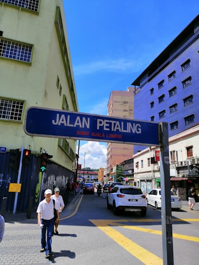 A street scene in an urban area with a street sign reading 'Jalan Petaling 50000 Kuala Lumpur'. The road is lined with buildings painted in various colors, including a lime green and a striking purple. There are vehicles on the street, including cars and a motorbike, along with pedestrians on the sidewalk. The sky is clear and blue.