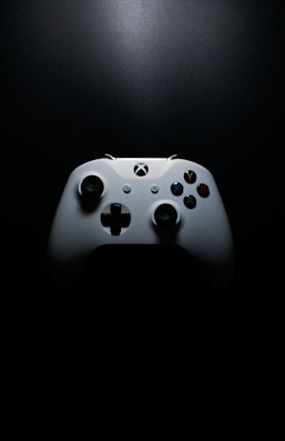 A white gaming controller with black buttons and colorful accents, placed on a dark background. The lighting creates a dramatic effect, highlighting the contours and buttons of the controller.