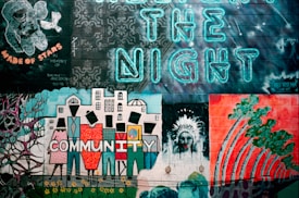A vibrant mural on a brick wall features various elements, including an astronaut, intricate floral patterns, stylized buildings, and abstract human figures. The word 'COMMUNITY' is boldly painted with each letter representing a person. The phrase 'CREATE THE NIGHT' is illuminated in neon blue letters above the artwork. There's a tribute to a person named Ralph Anderson, signed 'Made of Stars'.