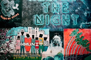 A vibrant mural on a brick wall features various elements, including an astronaut, intricate floral patterns, stylized buildings, and abstract human figures. The word 'COMMUNITY' is boldly painted with each letter representing a person. The phrase 'CREATE THE NIGHT' is illuminated in neon blue letters above the artwork. There's a tribute to a person named Ralph Anderson, signed 'Made of Stars'.