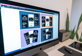 A computer monitor displaying a design mock-up for a mobile payment application. The screen shows six design variations with different color schemes and layouts. A desk setup is visible in the background, including a keyboard, mouse, graphics tablet, and some plants.