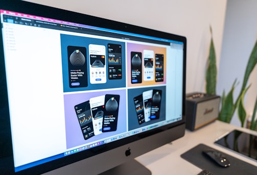 A computer monitor displaying a design mock-up for a mobile payment application. The screen shows six design variations with different color schemes and layouts. A desk setup is visible in the background, including a keyboard, mouse, graphics tablet, and some plants.