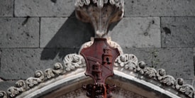 An ornate architectural detail featuring a carved stone emblem with two towers, set against a textured grey brick wall. The emblem is reddish in color, with intricate white floral stone carvings surrounding the archway.