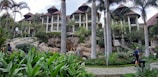 A luxurious resort featuring several multi-story buildings with thatched roofs, surrounded by lush greenery and tall palm trees. A stone pathway meanders through well-maintained gardens with vibrant tropical plants. People with luggage can be seen walking along the path, adding a sense of travel and exploration.