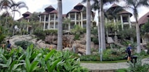 A luxurious resort featuring several multi-story buildings with thatched roofs, surrounded by lush greenery and tall palm trees. A stone pathway meanders through well-maintained gardens with vibrant tropical plants. People with luggage can be seen walking along the path, adding a sense of travel and exploration.