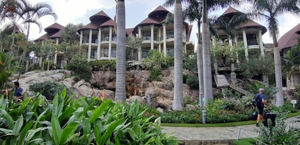 A luxurious resort featuring several multi-story buildings with thatched roofs, surrounded by lush greenery and tall palm trees. A stone pathway meanders through well-maintained gardens with vibrant tropical plants. People with luggage can be seen walking along the path, adding a sense of travel and exploration.