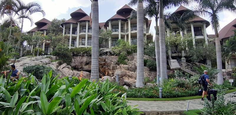 A luxurious resort featuring several multi-story buildings with thatched roofs, surrounded by lush greenery and tall palm trees. A stone pathway meanders through well-maintained gardens with vibrant tropical plants. People with luggage can be seen walking along the path, adding a sense of travel and exploration.