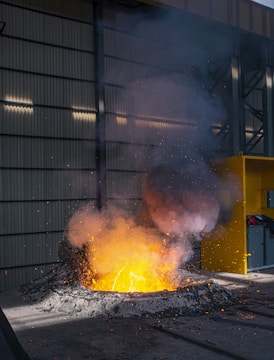 A molten metal pool emits a bright orange and yellow glow, surrounded by dark debris and scattered sparks. Smoke rises above the intense heat, while the scene is set in an industrial facility with metal siding and a yellow safety barrier.