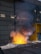 A molten metal pool emits a bright orange and yellow glow, surrounded by dark debris and scattered sparks. Smoke rises above the intense heat, while the scene is set in an industrial facility with metal siding and a yellow safety barrier.