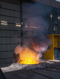 A molten metal pool emits a bright orange and yellow glow, surrounded by dark debris and scattered sparks. Smoke rises above the intense heat, while the scene is set in an industrial facility with metal siding and a yellow safety barrier.