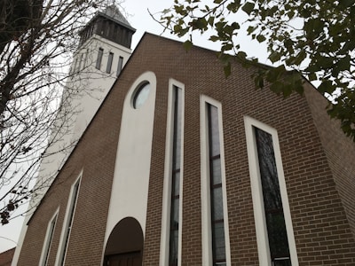 A modern-looking church building with a tall, white bell tower. The facade of the church features large, narrow windows and a brown brick exterior, accented with white columns. Leafless branches frame the view from both sides, suggesting a late autumn or winter setting.