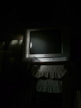 A dimly lit room features an older model CRT television set, with a silver-gray frame and a black screen. Below the TV, there are two fabric pieces with ruffled edges, possibly decorative covers or dust ruffles. The overall light is minimal, creating a shadowy ambiance.
