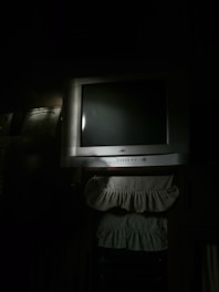 A dimly lit room features an older model CRT television set, with a silver-gray frame and a black screen. Below the TV, there are two fabric pieces with ruffled edges, possibly decorative covers or dust ruffles. The overall light is minimal, creating a shadowy ambiance.