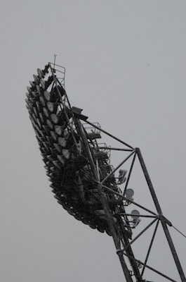An array of large, industrial floodlights mounted on a tall metal structure against a gray sky. The lights are arranged in multiple rows, supported by a complex framework of metal beams and platforms.