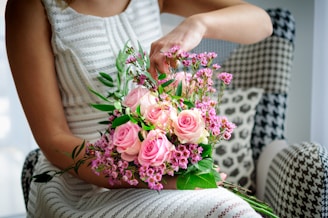 A person arranges a bouquet with pink roses and small purple flowers while seated on a patterned chair. The arrangement of flowers is vibrant and lush, with green leaves adding contrast. The person is wearing a white textured dress that complements the soft colors of the bouquet.
