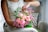 A person arranges a bouquet with pink roses and small purple flowers while seated on a patterned chair. The arrangement of flowers is vibrant and lush, with green leaves adding contrast. The person is wearing a white textured dress that complements the soft colors of the bouquet.