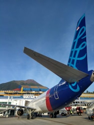 An airplane is parked on the tarmac in front of a terminal building with the words 'Bandar Udara Sultan'. The tail of the aircraft displays a blue graphic design with a flag at the top. A mountain and clear blue sky are visible in the background. Ground service personnel and vehicles can be seen near the airplane.