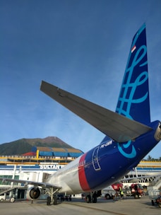 An airplane is parked on the tarmac in front of a terminal building with the words 'Bandar Udara Sultan'. The tail of the aircraft displays a blue graphic design with a flag at the top. A mountain and clear blue sky are visible in the background. Ground service personnel and vehicles can be seen near the airplane.