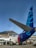 An airplane is parked on the tarmac in front of a terminal building with the words 'Bandar Udara Sultan'. The tail of the aircraft displays a blue graphic design with a flag at the top. A mountain and clear blue sky are visible in the background. Ground service personnel and vehicles can be seen near the airplane.