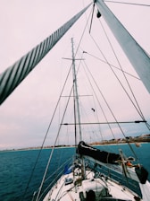 A sailboat is viewed from its deck looking up toward the tall mast and rigging. The sky is overcast, and the horizon shows a coastline with buildings in the distance. The ocean appears calm and the boat is equipped with various nautical items.