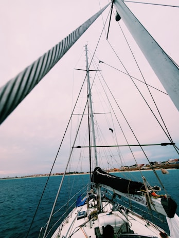 A sailboat is viewed from its deck looking up toward the tall mast and rigging. The sky is overcast, and the horizon shows a coastline with buildings in the distance. The ocean appears calm and the boat is equipped with various nautical items.