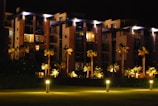 A modern apartment building at night with illuminated walkways and palm trees lining the path. The building features large balconies and is surrounded by well-maintained greenery. Soft lighting creates a warm, inviting atmosphere.
