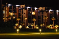 A modern apartment building at night with illuminated walkways and palm trees lining the path. The building features large balconies and is surrounded by well-maintained greenery. Soft lighting creates a warm, inviting atmosphere.