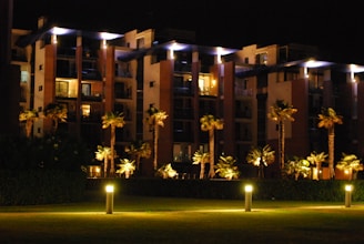 A modern apartment building at night with illuminated walkways and palm trees lining the path. The building features large balconies and is surrounded by well-maintained greenery. Soft lighting creates a warm, inviting atmosphere.