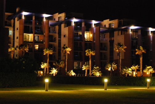 A modern apartment building at night with illuminated walkways and palm trees lining the path. The building features large balconies and is surrounded by well-maintained greenery. Soft lighting creates a warm, inviting atmosphere.