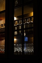 A dimly lit wooden cabinet showcases rows of bottled spirits on the top shelf and an assortment of elegant glassware on the bottom shelf. The lighting casts a warm, ambient glow, enhancing the rich tones of the wood and the reflections on the glass surfaces.