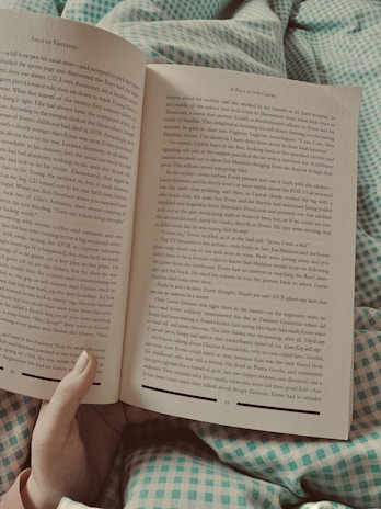An open book displaying two pages with text, held by a person whose hand is partially visible. The book rests on a green and white checkered fabric, possibly a blanket or bedspread. The surrounding lighting appears soft, suggesting a relaxed reading environment.