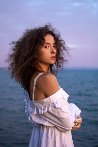A young woman with curly hair is wearing an off-the-shoulder white dress with tassel details. She is standing by the sea during a sunset, with the sky painted in soft pink and purple hues.