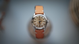 A vintage wristwatch with a round, metallic case and a worn-looking face. The watch features long, slim hour markers and small black hands. It is attached to a light brown leather strap, which adds a classic touch to the overall appearance.