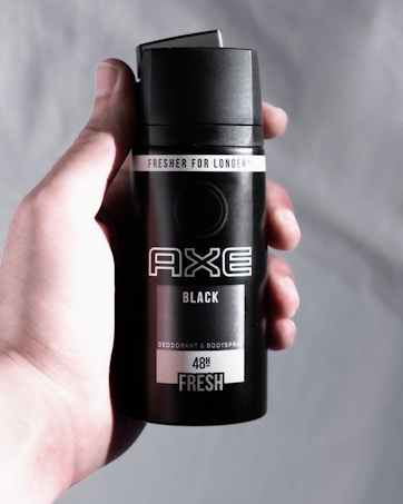 A hand holds a black canister of AXE deodorant and body spray with 'Fresher for Longer' and '48H Fresh' labels. The can is sleek and minimalist in design, featuring bold, white text.