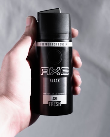 A hand holds a black canister of AXE deodorant and body spray with 'Fresher for Longer' and '48H Fresh' labels. The can is sleek and minimalist in design, featuring bold, white text.