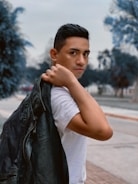 A person is holding a black leather jacket over their shoulder while wearing a white shirt. The background features an outdoor setting with blurred trees and a pathway.