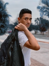A person is holding a black leather jacket over their shoulder while wearing a white shirt. The background features an outdoor setting with blurred trees and a pathway.