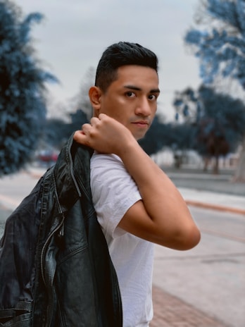 A person is holding a black leather jacket over their shoulder while wearing a white shirt. The background features an outdoor setting with blurred trees and a pathway.