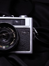 A vintage black and silver camera, likely made by Ricoh, is positioned against a dark fabric backdrop. The camera features a textured grip and a prominent lens with visible markings indicating lens specifications.