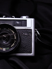 A vintage black and silver camera, likely made by Ricoh, is positioned against a dark fabric backdrop. The camera features a textured grip and a prominent lens with visible markings indicating lens specifications.